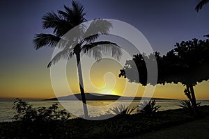 Dramatic photo of ocean, palm trees and Lanai from a beach on Maui at sunset.
