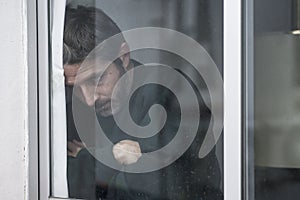 Dramatic portrait of attractive sad and depressed mid adult man looking through home window feeling worried and desperate
