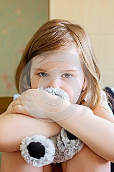 Dramatic portrait of little girl sitting on chair and cuddling teddy bear with sad face.