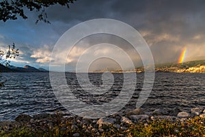 Dramatic storm clouds and rain passing over Okanagan Lake with view of a double rainbow above Naramata