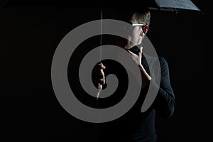 Dramatic studio portrait of a guy in black, holding an umbrella in sunglasses. On a gray background.