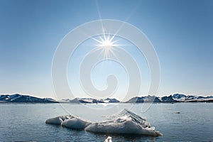 A dramatic sun burst over arctic mountains and a floating iceberg in foreground floats on calm waters