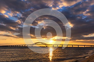 Dramatic sunset light and clouds over a long steel tied arch bridge. Fire Island Inlet Bridge