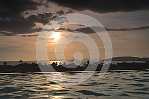 Dramatic tropical evening Sun in Thailand - view over roof top swimming pool in luxury hotel. Dark clouds over palms, sea