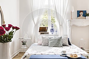 Drapes at window above bed with cushions in white bedroom interior with poster and roses. Real photo