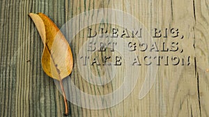 Dream BIG, Set GOALS, Take ACTION phrase. Written on wooden surface. yellowed leaf