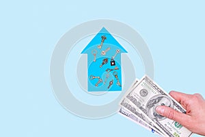 Dream home concept. Woman`s hand holds dollars over a model blue paper house and keys on a pastel background