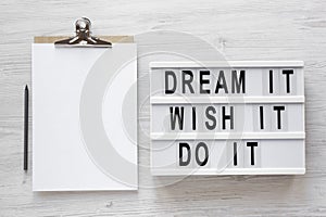 `Dream it, wish it, do it` words on a modern board, clipboard with blank sheet of paper on a white wooden surface, top view.