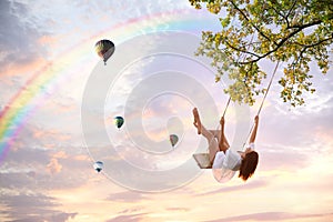 Dream world. Young woman swinging, hot air balloons in sunset sky on background