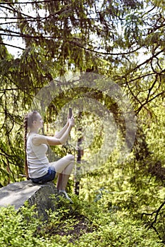 Dreaming beautiful girl sitting on a stone and making selfie on a smartphone surrounded by coniferous forest on a sunny day