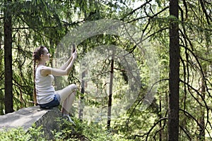 Dreaming beautiful girl sitting on a stone and making selfie on a smartphone surrounded by coniferous forest on a sunny day