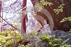 Dreaming beautiful girl sitting on a stone under a rock surrounded by forest on a sunny day