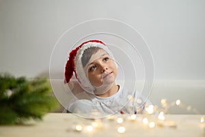 Dreaming inspired little kid boy in the christmas head with garland lights and fir-tree branch on the table