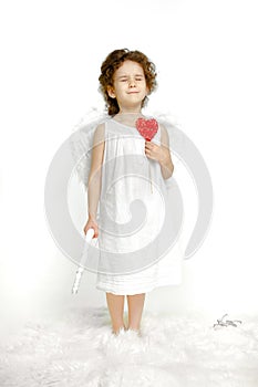 Dreaming little girl with curly hair, in white dress and wings, over white background. Angel holding a red heart.