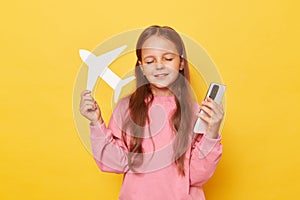 Dreaming little girl wearing pink sweatshirt holding white paper plane and phone isolated over yellow background thinking about