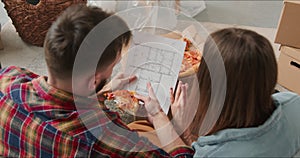 Dreaming of new home. Young happy couple eat pizza on the floor looking at new apartment plan blueprint planning future.