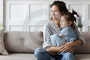 Dreamy affectionate curly mother cuddling small preschool daughter, looking away.