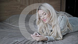 Dreamy Beautiful blonde woman lying on the bed with smart phone. Girl smiles, good mood. Blogging, browsing internet