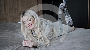 Dreamy Beautiful blonde woman lying on the bed with smart phone. Girl smiles, good mood. Blogging, browsing internet