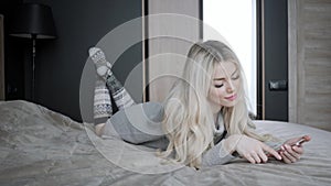 Dreamy Beautiful blonde woman lying on the bed with smart phone. Girl smiles, good mood. Blogging, browsing internet