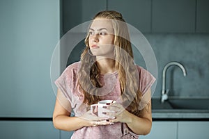 Dreamy beautiful young woman with cup of hot coffee in kitchen in morning. Soft pink pajamas