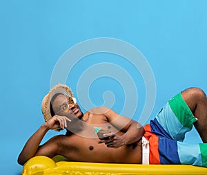 Dreamy black man sunbathing on inflatable lilo, drinking tropical cocktail from coconut shell on blue background