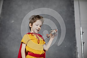 Dreamy boy in a red cape plays with an airplane toy on a gray artistic background