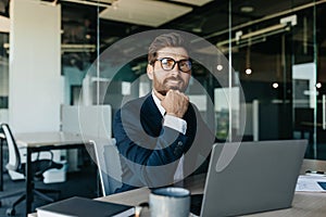 Dreamy businessman sitting at workplace in office and thinking of new business strategy, looking aside and smiling