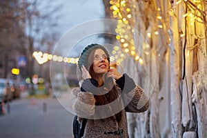 Dreamy caucasian woman smiling in the evening street. City lights. Winter casual clothes. Festive city.