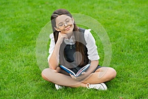 dreamy child in glasses reading book sitting on green grass
