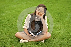 dreamy child in glasses reading book sitting on green grass