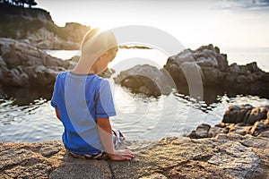 Dreamy child on sea. Little boy sits on rocky beach at sunset and look at surface of waves sea water