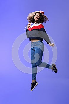 Dreamy and femenine cute girl with curly hair in winter beanie, jeans and sweatshirt jumping over blue background with