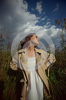 Dreamy gentle woman in a beige raincoat in nature. Romantic young woman in thick tall grass in field. Natural beauty, enjoyment of