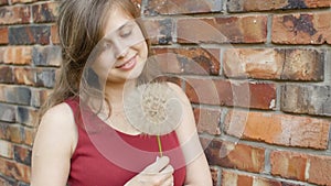 Dreamy girl with fluffy big dandelion on the background of the red brick wall, beautiful young woman smilng and flirting, female e