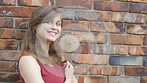 Dreamy girl with fluffy big dandelion on the background of the red brick wall, beautiful young woman smilng and flirting, female e