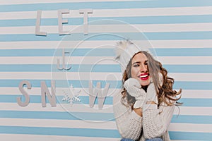 Dreamy girl in winter knitted outfit posing with pleasure on striped background. Brunette curly lady in woolen mittens