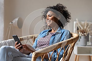 Dreamy happy African American woman holding smartphone, looking in distance