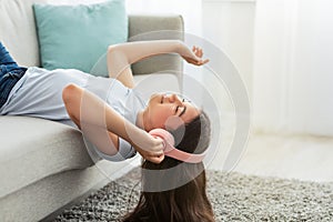 Dreamy Indian teen girl in wireless headphones listening to music or audio book, lying on couch at home, free space