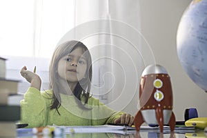 Dreamy little girl with pencil is doing homework while dreaming of a better world - Elementary schooler girl at home while drawing