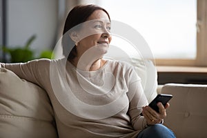 Dreamy middle aged woman resting on couch, holding phone