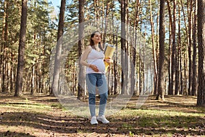 Dreamy pensive pregnant woman wearing casual clothing holding karemat while standing in spring wood and enjoying nature taking