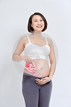 Dreamy pregnant woman holding baby booties near tummy on white