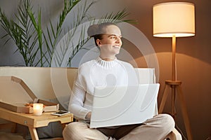 Dreamy smiling caucasian woman with bun hairstyle in white sweater looking at laptop screen sitting on sofa at home looking away