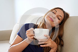 Dreamy smiling positive girl eyes closed holds cup with warm beverage imagining sitting on sofa