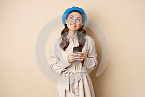 Dreamy smiling woman in stylish trenchcoat, looking up fantasizing while shopping on mobile phone app, using smartphone