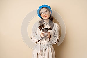 Dreamy smiling woman in stylish trenchcoat, looking up fantasizing while shopping on mobile phone app, using smartphone