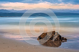 Dreamy waves breaking in slow motion at Plettenberg Bay beach at sunset, with hills in the distance and a rock in the foreground