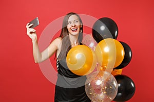 Dreamy woman in black dress celebrating holding air balloons doing taking selfie shot on mobile phone isolated on red