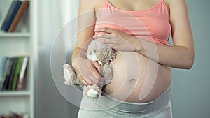 Dreamy woman with pregnant belly hugging toy bear tenderly, surrogate mother